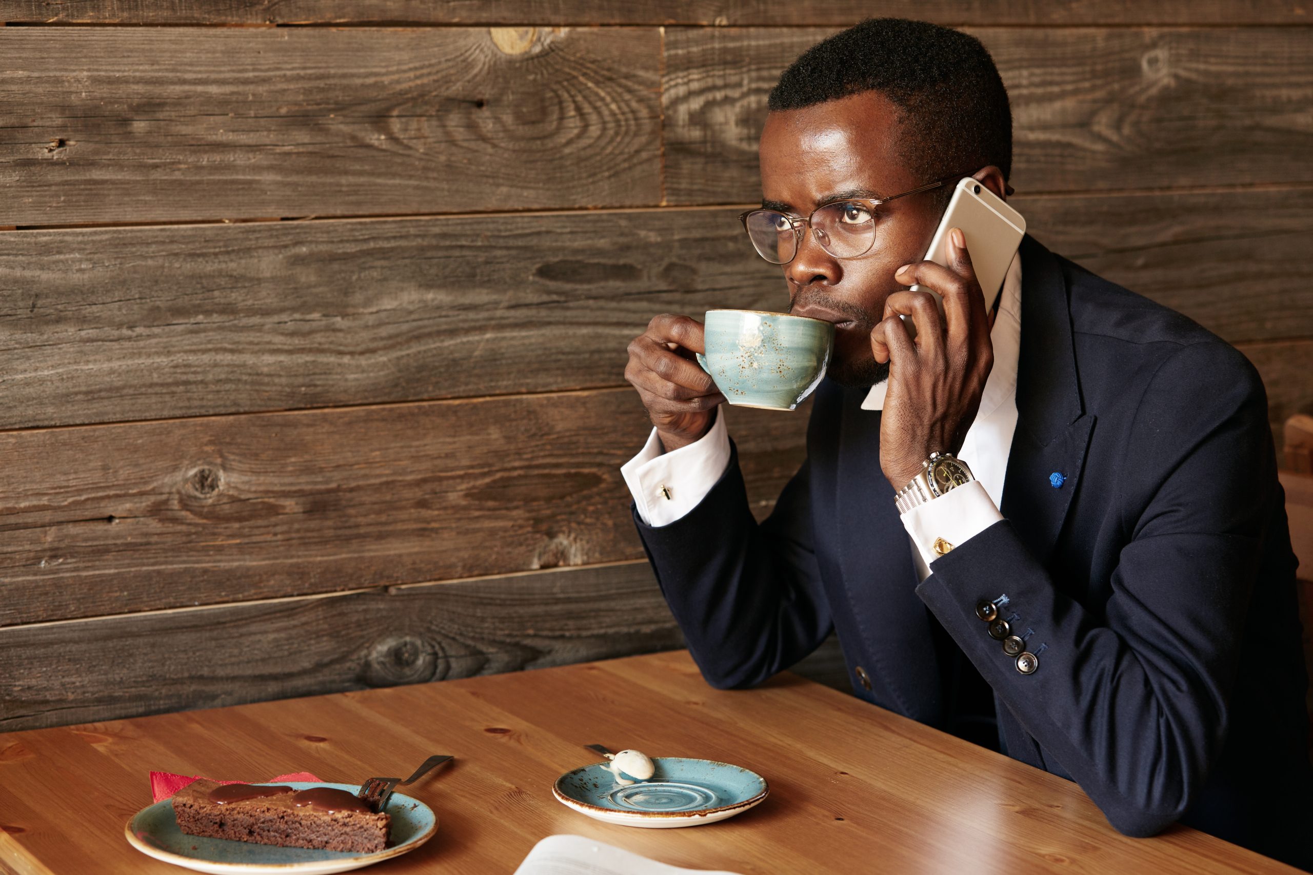 Successful young African CEO talking on the phone with serious and concentrated expression. Black student drinking cappuccino and having dessert, making phone calls while relaxing at the cafe