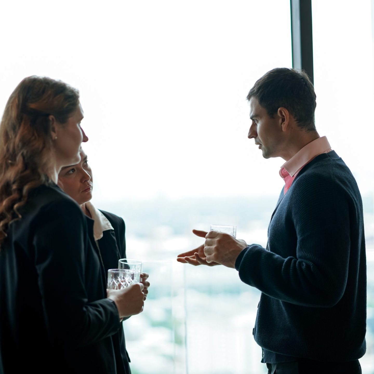 A group of people are standing in a room, one of them is talking to another. The man is wearing a blue sweater and holding a glass of water. The woman is wearing a black jacket