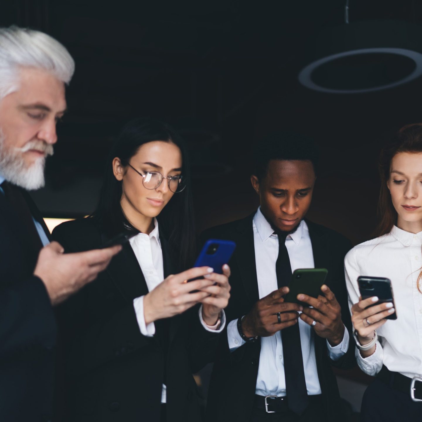 Group of multiracial team partners wearing formal clothes standing close to each other and browsing smartphones in darkly lit space