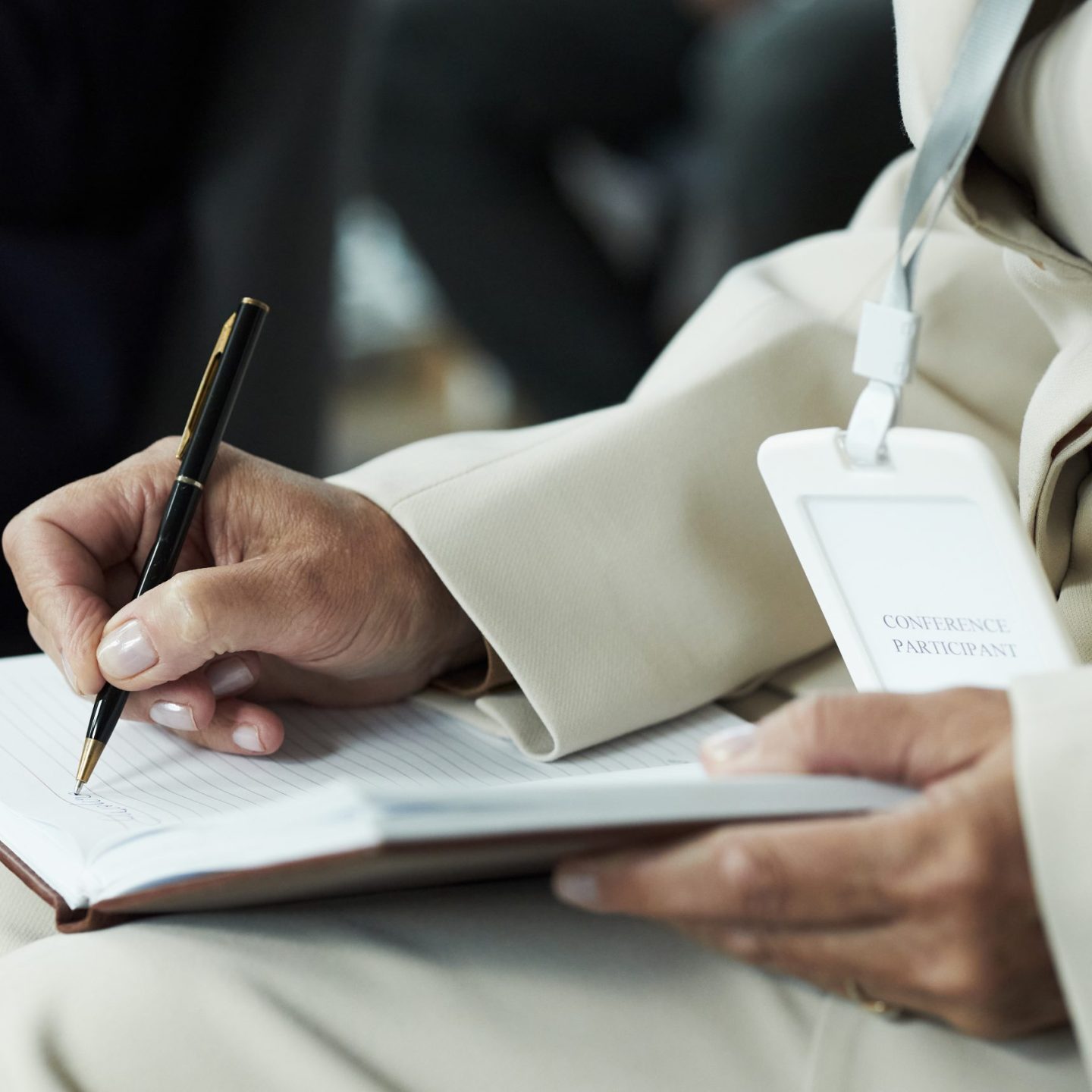 Middle aged unrecognizable woman writing in notebook while attending conference, holding pen in right hand and wearing visible name badge, focusing on taking notes during event