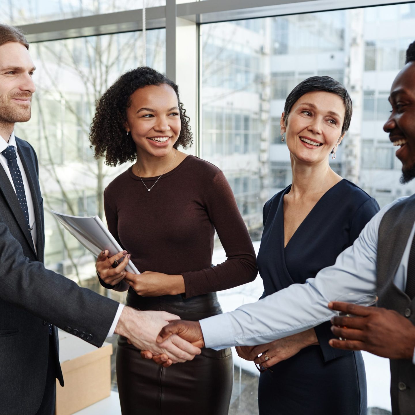 Waist up portrait of smiling business partners shaking hands in office standing with drinks and celebrating deal