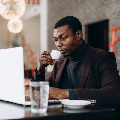 Sixth Hive Investor Young Black man in smart casual attire sipping coffee while focused on laptop work at a modern cafe table during daytime