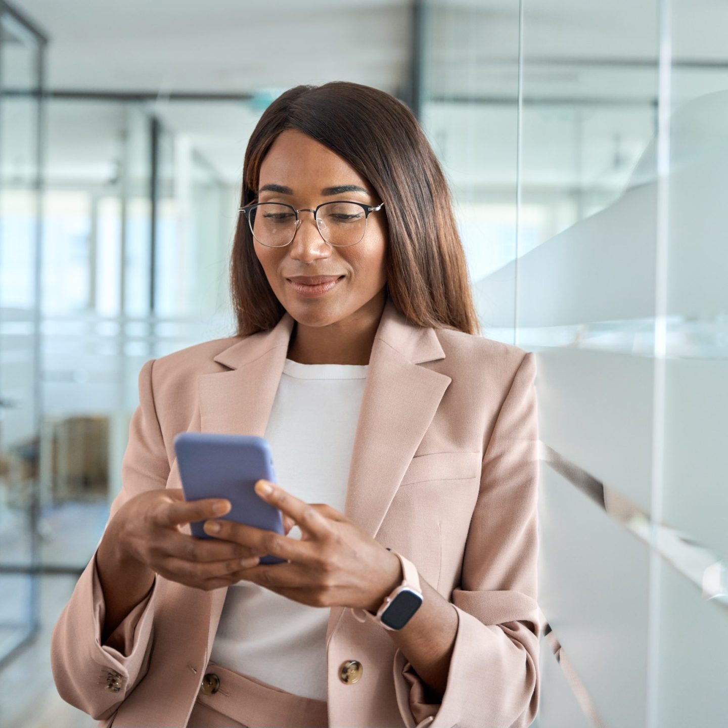 Young busy professional African American business woman company executive manager or entrepreneur wearing suit holding cellphone using mobile phone standing in office working on smartphone.